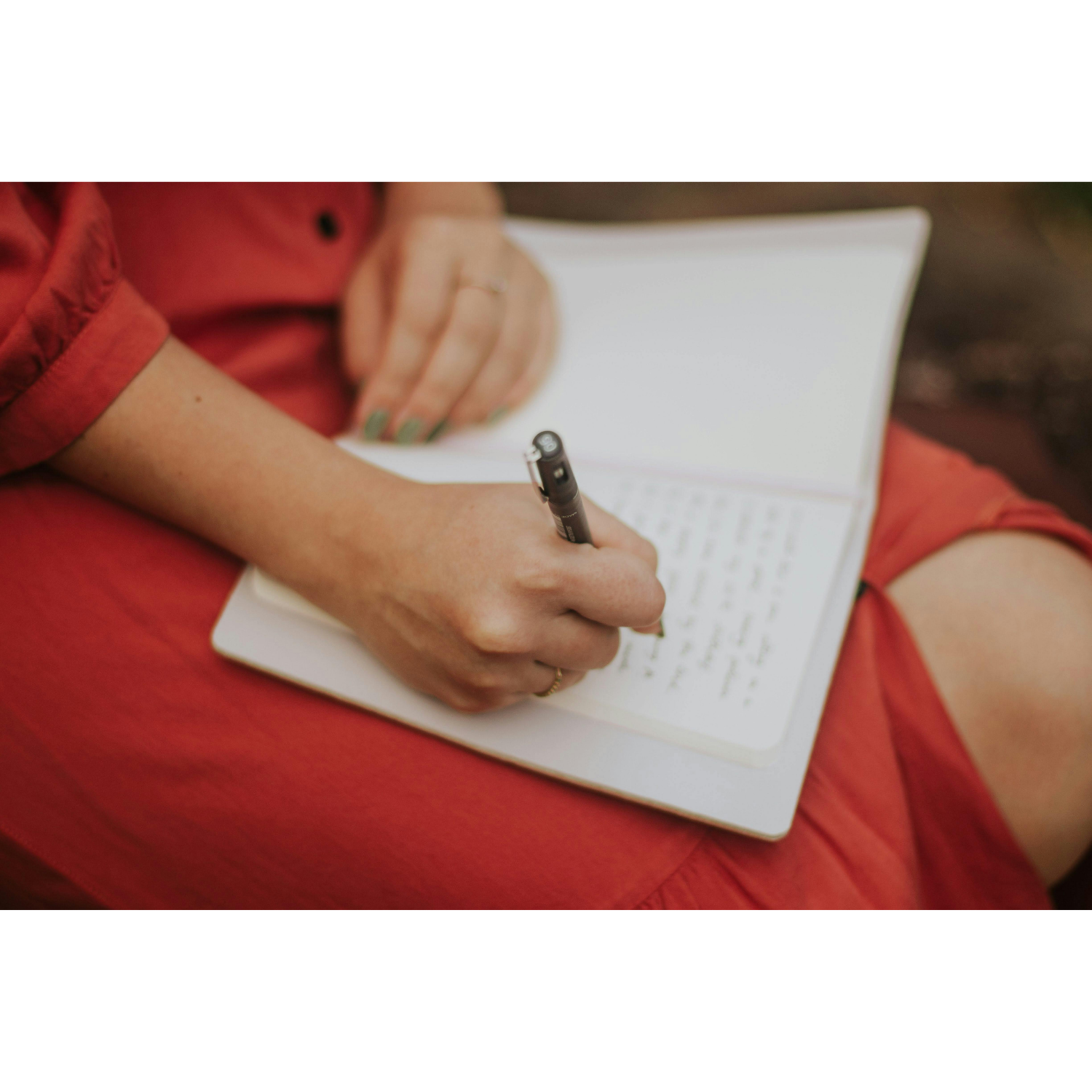 A woman in a red dress writing in an open notebook on her lap, with a pen in hand and a delicate gold ring on her finger