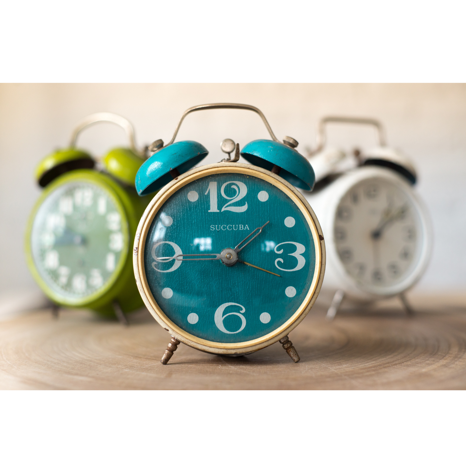 Three vintage twin-bell alarm clocks on a wooden surface, with a teal clock in sharp focus and green and white clocks softly blurred behind it