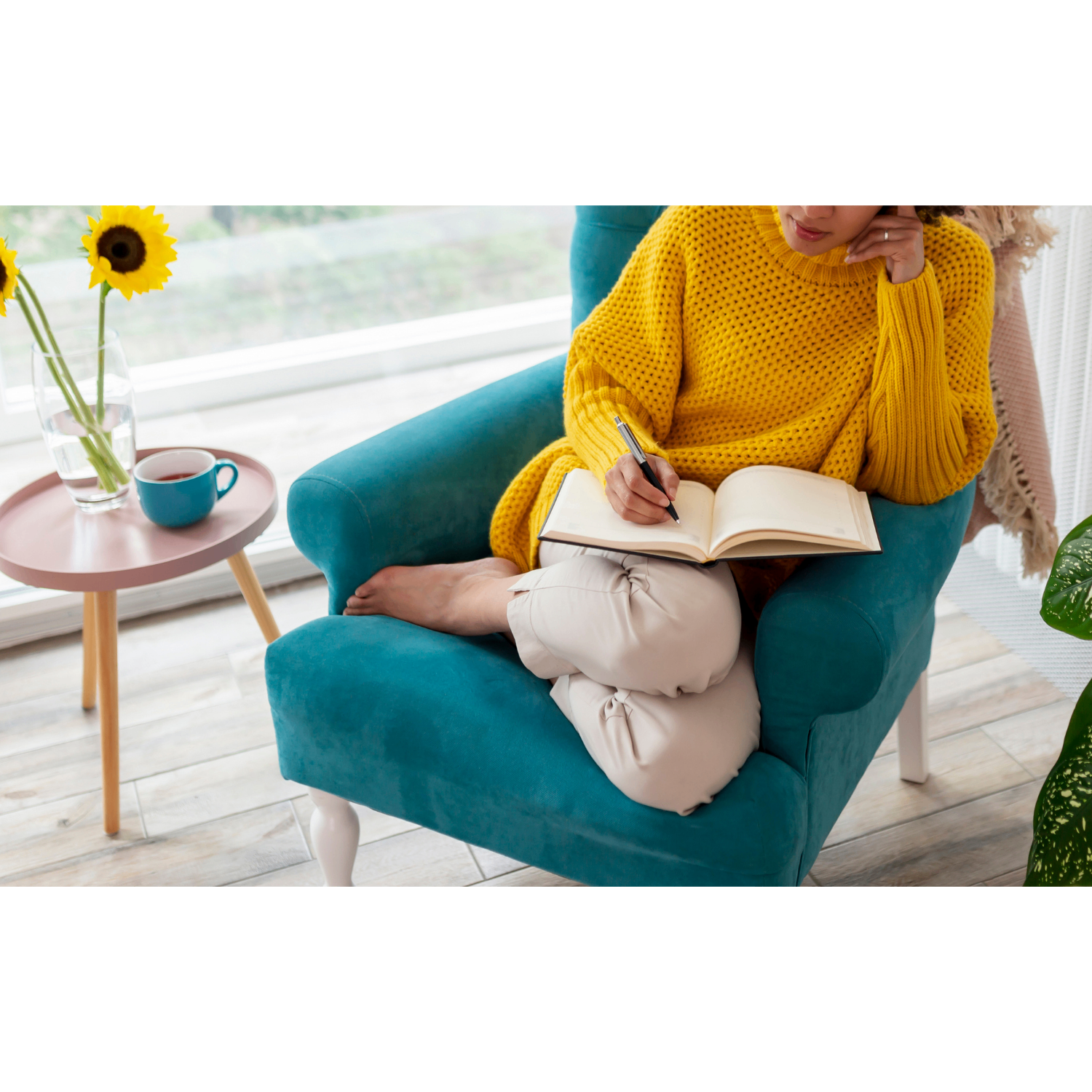 A woman in a yellow knit sweater curled up in a teal armchair, writing in a journal beside a small side table with sunflowers and a mug of tea